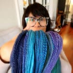 A person with short dark hair and glasses holds up several skeins of multicolored blue, teal, and purple yarn in a cozy, well-lit room with wooden floors and light furniture.