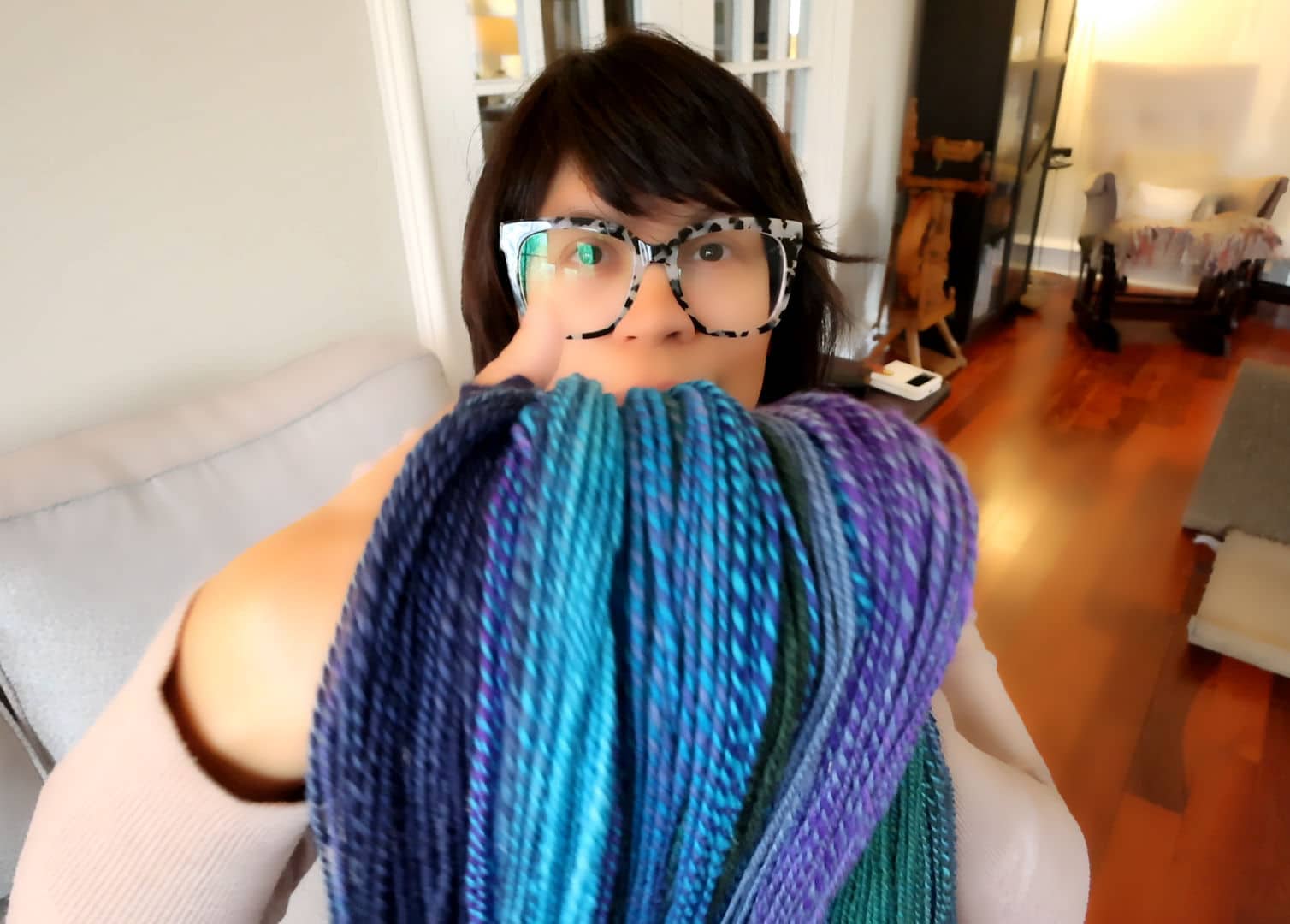 A person with short dark hair and glasses holds up several skeins of multicolored blue, teal, and purple yarn in a cozy, well-lit room with wooden floors and light furniture.