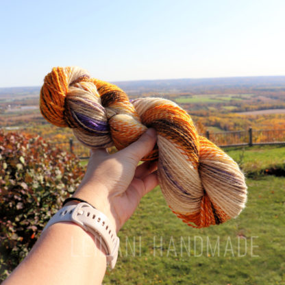 A hand wearing a beige smartwatch holds a skein of orange, cream, brown, and purple yarn. The background shows a scenic countryside landscape with fields, trees, and a distant horizon under a clear blue sky. Text reads “LEILANI HANDMADE.”.