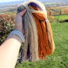 A hand holds an open skein of yarn in cream, orange, purple, and brown colors outdoors. The handspun yarn is called, "Lilacs in Autumn". The sunny background features green grass, shrubs, a fence, and a distant view of rolling hills and trees under a clear sky.