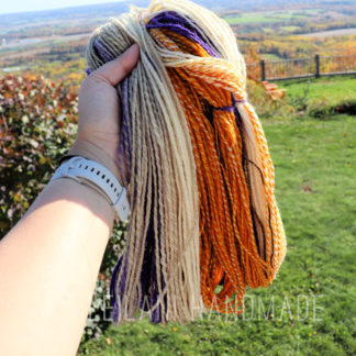 A hand holds an open skein of yarn in cream, orange, purple, and brown colors outdoors. The handspun yarn is called, "Lilacs in Autumn". The sunny background features green grass, shrubs, a fence, and a distant view of rolling hills and trees under a clear sky.