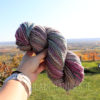 A hand wearing a beige watch holds up a skein of multicolored yarn with pink, blue, and gray hues. The background shows a scenic, sunlit landscape with rolling hills, trees, and a clear sky. The words “LEILANI HANDMADE” are faintly visible.