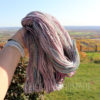 A hand wearing a beige watch holds up a skein of pastel purple, pink, and blue twisted yarn outdoors. The background shows green fields, trees, and a distant landscape under a clear sky on a sunny day.