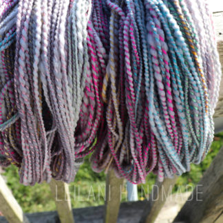 A large spiral ply handspun yarn, twisted and hung on a wooden fence. The yarn features shades of purple, pink, blue, and grey tones. Sunlight highlights the vibrant colors, and the words "LEILANI HANDMADE" appear at the bottom.