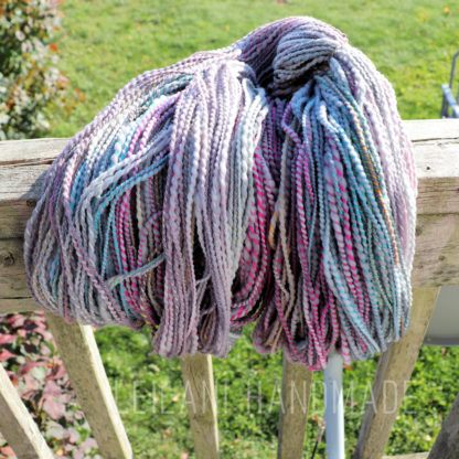 A skein of handspun yarn with pastel shades of pink, blue, grey, and hints of yellow hangs draped over a weathered wooden railing outdoors on a sunny day. Grass and garden plants are visible in the blurred background.