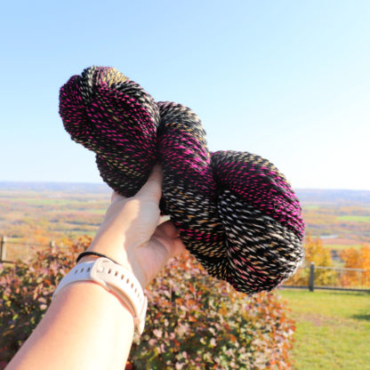 A hand wearing a pale smartwatch holds up a skein of twisted yarn in shades of black, white, pink, and green. In the background, there’s a blurred autumn landscape with fields, trees in fall colors, and a blue sky.