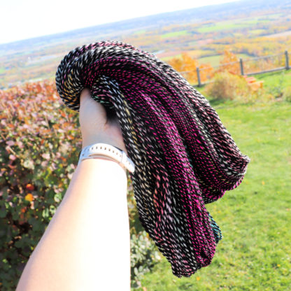 A hand wearing a white watch holds up a hank of twisted yarn in black, white, and magenta colors. The background shows a grassy yard, autumn bushes, a wooden fence, and a distant landscape under a bright sky.