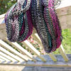 Coiled yarn with black, white, magenta, and teal stripes hang over a weathered wooden railing in bright sunlight. The background shows green grass and blurred foliage. Shadows from the railing create lines on the wooden deck below.