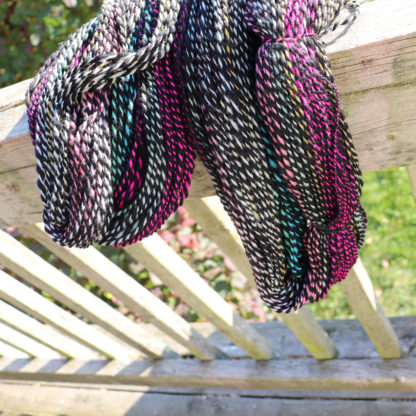 Coiled yarn with black, white, magenta, and teal stripes hang over a weathered wooden railing in bright sunlight. The background shows green grass and blurred foliage. Shadows from the railing create lines on the wooden deck below.