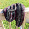 A skein of hand-dyed, twisted yarn in shades of black, white, magenta, and teal drapes over a weathered wooden railing outdoors, with bright green grass and sunlight in the background.