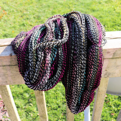 A skein of hand-dyed, twisted yarn in shades of black, white, magenta, and teal drapes over a weathered wooden railing outdoors, with bright green grass and sunlight in the background.