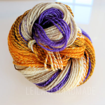 A skein of twisted yarn in cream, orange, yellow, and purple hues is coiled on a glossy white surface. The yarn appears thick and soft, and the colors are vibrant and variegated. The text “LEILANI HANDMADE” is faintly visible at the bottom.