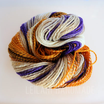 A skein of yarn twisted into a coil, featuring a mix of cream, orange, brown, and purple strands. The colors are blended in a barber pole pattern, and the text “LEILANI HANDMADE” is faintly visible at the bottom of the image. The handspun yarn is entitled, "Lilacs in Autumn".