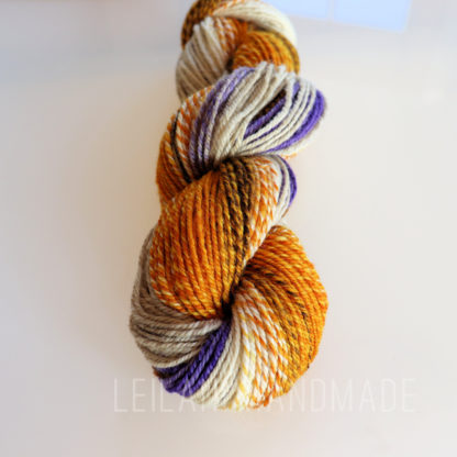 A twisted skein of yarn with colors including golden yellow, orange, cream, brown, and purple sits on a smooth, pale surface. The yarn appears soft and has a subtle sheen, with the colors blending and contrasting in a variegated pattern.