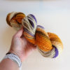 A hand holds a skein of yarn with a blend of orange, yellow, cream, brown, and purple colors. The person wears a light-colored smartwatch. The background is plain and light, and faint text reads "LEILANI HANDMADE" near the bottom.