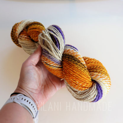 A hand holds a skein of yarn with a blend of orange, yellow, cream, brown, and purple colors. The person wears a light-colored smartwatch. The background is plain and light, and faint text reads "LEILANI HANDMADE" near the bottom.
