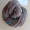 A skein of handspun yarn in shades of gray with subtle hints of pink, green, and yellow twists, coiled into a neat bundle on a reflective white surface. Sunlight and a window are faintly reflected in the background.