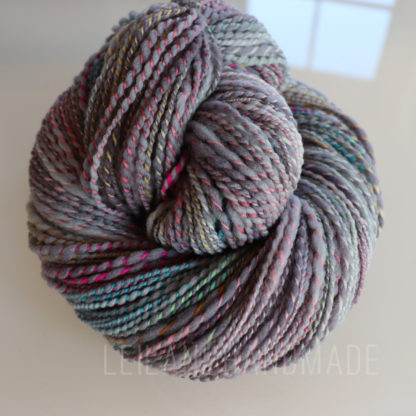 A skein of handspun yarn in shades of gray with subtle hints of pink, green, and yellow twists, coiled into a neat bundle on a reflective white surface. Sunlight and a window are faintly reflected in the background.