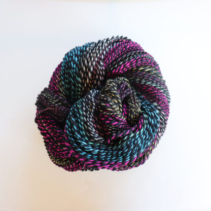 A skein of twisted yarn arranged in a round shape, featuring variegated colors of black, blue, pink, white, and green, set against a plain white background. The fiber appears thick, soft, and slightly glossy.