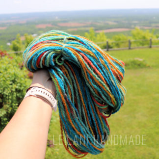 A hand wearing a light-colored watch holds a skein of colorful yarn outdoors, with a blurred green lawn, trees, and distant fields visible in the background. The text "LEILANI HANDMADE" is overlaid at the bottom.