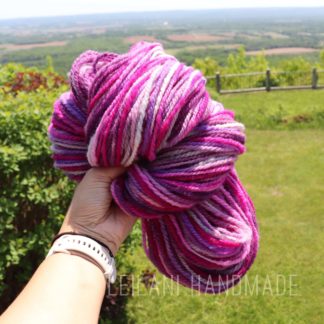 A hand holding a skein of pink, purple, and white yarn outdoors, with a scenic green landscape and distant horizon in the background. The text “LEILANI HANDMADE” is faintly visible at the bottom.