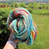 A hand holds a colorful skein of yarn outdoors. The yarn features vibrant shades of blue, yellow, orange, red, and green. In the background, there’s a grassy lawn, trees, a wooden fence, and rolling hills under a partly cloudy sky.
