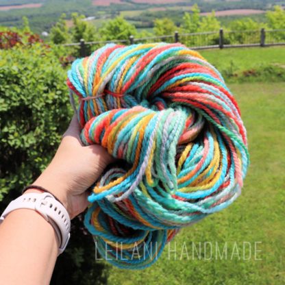 A hand holds a skein of colorful, multicolored yarn in an outdoor setting with green grass, bushes, and a scenic landscape in the background. The words "LEILANI HANDMADE" appear at the bottom.