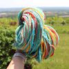 A hand holds up a skein of multicolored yarn in bright shades of yellow, blue, orange, red, and white. The background shows a lush green yard, trees, and a hazy view of fields and sky. The words "LEILANI HANDMADE" are faintly overlaid.