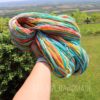 A hand holds a skein of colorful yarn with shades of blue, yellow, orange, and white, against a vibrant green field and distant hills under a bright sky. The text "LEILANI HANDMADE" is faintly visible at the bottom of the image.