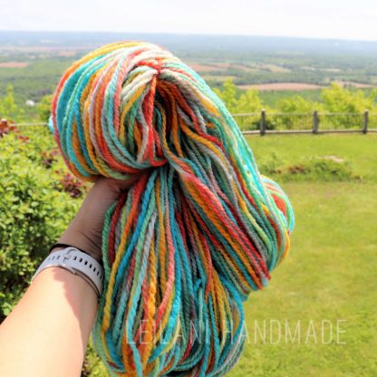 A hand holds a skein of colorful yarn—blue, yellow, orange, and white—against a bright green grassy landscape with trees, fields, and a distant horizon under a clear sky. The person wears a white watch.