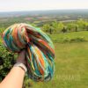 A hand holds a large skein of colorful, multicolored yarn outdoors. In the background, there is a lush green lawn, a wooden fence, trees, and a scenic view of fields and forests stretching to the horizon under a clear sky.