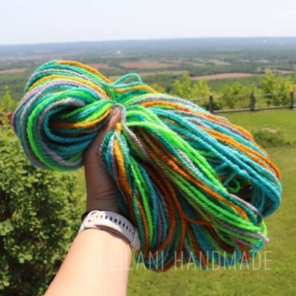 A hand holds a bundle of thick, multicolored yarn in green, blue, orange, yellow, and gray strands, with a scenic landscape of distant fields, trees, and blue sky in the background. The words "LEILANI HANDMADE" are faintly visible at the bottom.