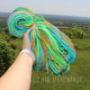 A hand holding a bundle of colorful yarn in shades of blue, green, yellow, and orange, with a scenic view of green fields and distant hills in the background. Text reads "LEILANI HANDMADE".