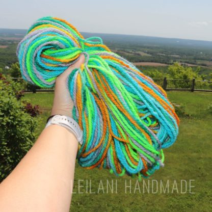 A hand holding a bundle of colorful yarn in shades of blue, green, yellow, and orange, with a scenic view of green fields and distant hills in the background. Text reads "LEILANI HANDMADE".