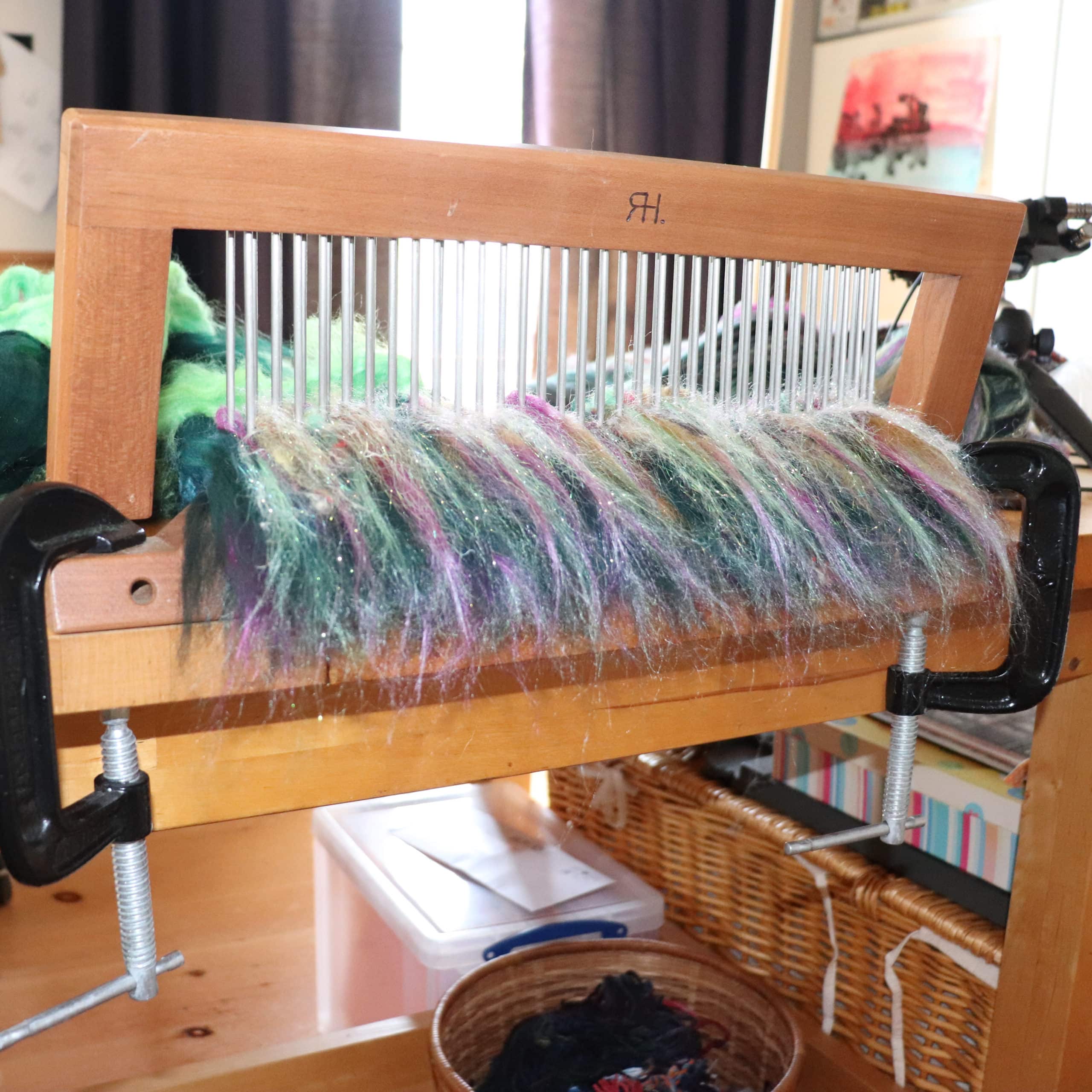 A wooden blending board with metal rods holds various colored fibers, including green, purple, and white, clamped to a table. The background shows baskets, a clear plastic storage box, and art on the walls.