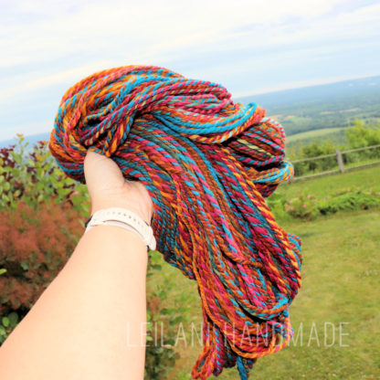 A hand holds up a thick skein of colorful, twisted yarn (red, blue, yellow, teal) against a backdrop of green grass, bushes, a fence, and a distant view of trees and blue sky. The words “LEILANIHANDMADE” appear at the bottom.