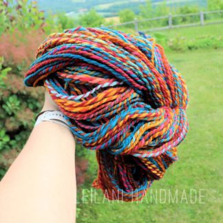 A hand holds a large skein of multicolored, twisted yarn, handspun and called "Autumn Vibes" featuring vibrant red, orange, blue, yellow, and black hues. The background shows a lush green lawn, shrubs, and distant hills under a bright sky. Text reads “LEILANI HANDMADE”.