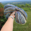 A hand wearing a white watch holds up a skein of multicolored yarn with shades of blue, tan, cream, and brown against a lush green landscape with trees, grass, distant hills, and a wooden fence. "LEILANI HANDMADE" is faintly visible in the foreground.