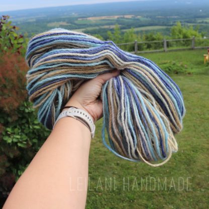 A hand wearing a white watch holds up a skein of multicolored yarn with shades of blue, tan, cream, and brown against a lush green landscape with trees, grass, distant hills, and a wooden fence. "LEILANI HANDMADE" is faintly visible in the foreground.