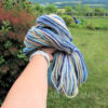 A hand holds a large skein of multicolored yarn called "Beach House" in blue, tan, cream, and gray tones outdoors. Lush green grass, bushes, and a wooden fence with scenic hills under a cloudy sky appear in the background. “LEILANI HANDMADE” is overlaid on the image.
