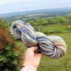 A hand holds a skein of multicolored yarn with shades of blue, beige, brown, and white. In the background, there is a lush green lawn, a wooden fence, trees, and a scenic view of fields and hills under a partly cloudy sky. Text reads “LEILANI HANDMADE”.