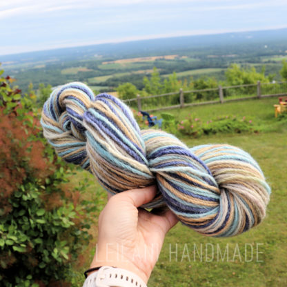 A hand holds a skein of multicolored yarn with shades of blue, beige, brown, and white. In the background, there is a lush green lawn, a wooden fence, trees, and a scenic view of fields and hills under a partly cloudy sky. Text reads “LEILANI HANDMADE”.