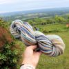 A hand holds a skein of multicolored yarn with shades of blue, beige, brown, and white. In the background, there is a lush green lawn, a wooden fence, trees, and a scenic view of fields and hills under a partly cloudy sky. Text reads “LEILANI HANDMADE”.