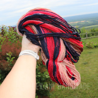 A hand wearing a white watch holds a skein of yarn in shades of red, navy blue, pink, and beige. This handspun yarn is called, "Red Sky at Night". The background shows a grassy lawn, bushes, a wooden fence, and a distant landscape under a blue sky. The text “LEILANI HANDMADE” appears at the bottom.