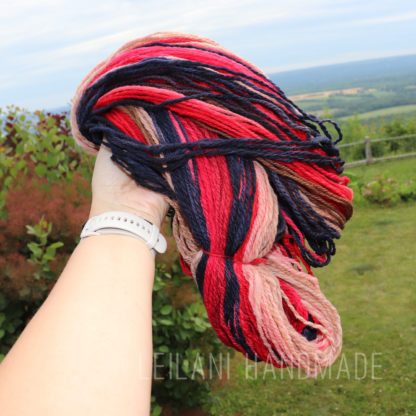 A hand wearing a white watch holds a skein of yarn in shades of red, navy blue, pink, and beige. This handspun yarn is called, "Red Sky at Night". The background shows a grassy lawn, bushes, a wooden fence, and a distant landscape under a blue sky. The text “LEILANI HANDMADE” appears at the bottom.