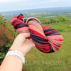 A hand holds a skein of multicolored yarn—pink, red, navy, and peach—against a scenic background of rolling green hills, trees, and a cloudy sky. The person wears a white watch. The words “LEILANI HANDMADE” appear at the bottom.