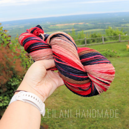 A hand holds a skein of multicolored yarn—pink, red, navy, and peach—against a scenic background of rolling green hills, trees, and a cloudy sky. The person wears a white watch. The words “LEILANI HANDMADE” appear at the bottom.