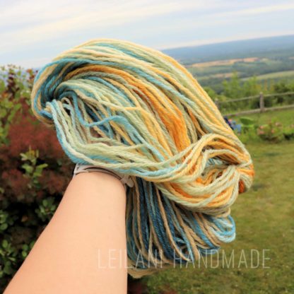 A hand holds a skein of multicolored yarn with shades of blue, cream, yellow, and orange. The background shows a green lawn, trees, and a distant landscape under a cloudy sky. The watermark reads “LEILANI HANDMADE”.