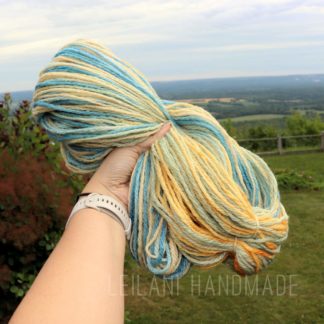 A hand holds a skein of handspun yarn in soft blue, yellow, and cream shades, called "Beachy Keen". The background shows a grassy yard, wooden fence, trees, and a distant landscape under a partly cloudy sky. “LEILANI HANDMADE” is faintly visible on the image.