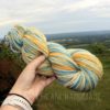 A hand holds a skein of thick, multicolored yarn in shades of yellow, blue, and light green. The background features a lush, green landscape and an expansive, cloudy sky. The text "LEILANI HANDMADE" is faintly visible at the bottom of the image.